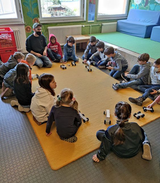 Kinder sitzen im Kreis auf einem gelben Teppich und bauen kleine Fahrzeuge aus LEGO oder Bausteinen.