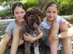 Zwei Kinder sitzen draußen und halten einen flauschigen Hund zwischen sich, beide lächeln in die Kamera.