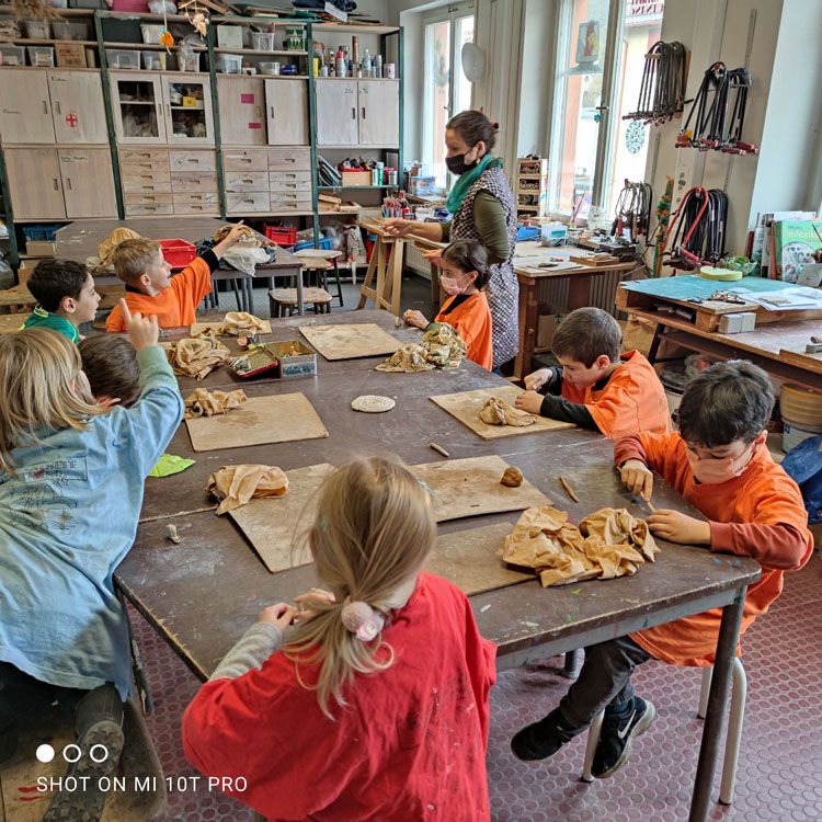 Kinder sitzen in einer Werkstatt an einem Tisch und arbeiten mit Ton, im Hintergrund eine Betreuerin.