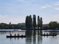Zwei Kanus mit Kindern auf einem See, im Hintergrund Bäume und Stadtgebäude.